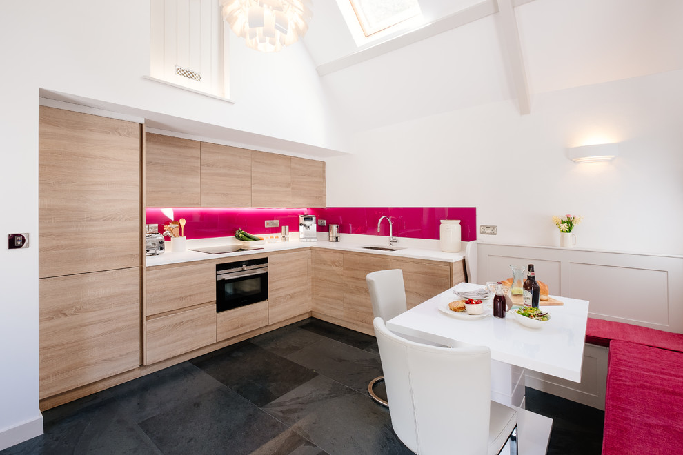 This is an example of a country l-shaped eat-in kitchen in Cornwall with an undermount sink, flat-panel cabinets, light wood cabinets, pink splashback, glass sheet splashback, no island, black floor and white benchtop.