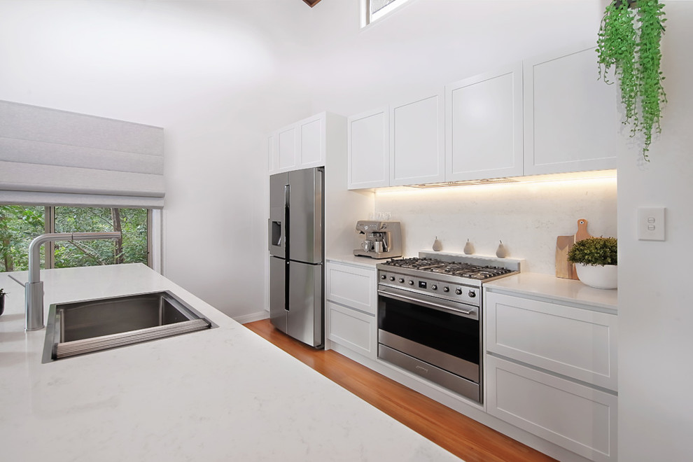 Inspiration for a large modern single-wall open plan kitchen in Sydney with a drop-in sink, recessed-panel cabinets, yellow cabinets, quartz benchtops, white splashback, glass sheet splashback, stainless steel appliances, light hardwood floors, with island, brown floor and white benchtop.