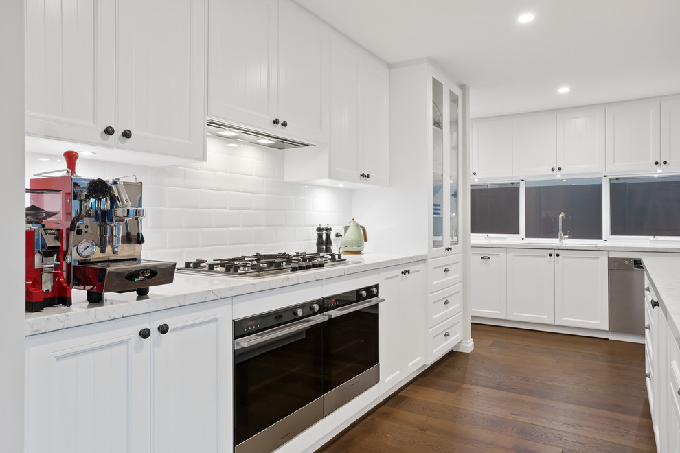 This is an example of a beach style galley open plan kitchen in Perth with a double-bowl sink, shaker cabinets, white cabinets, solid surface benchtops, white splashback, subway tile splashback, black appliances, medium hardwood floors, with island and white benchtop.