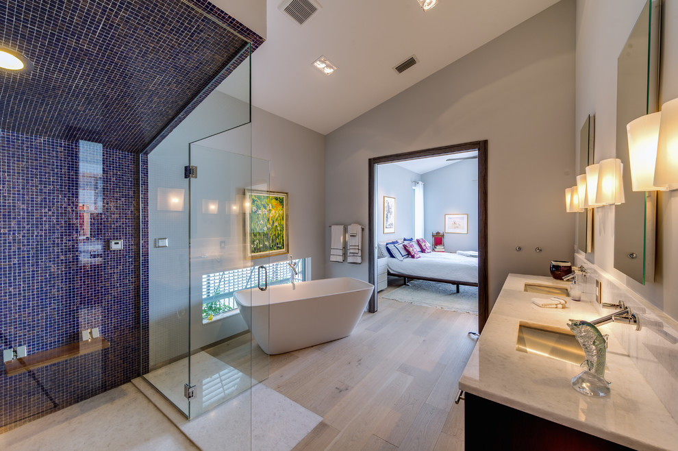 Photo of an expansive contemporary bathroom in Other with flat-panel cabinets, brown cabinets, a freestanding tub, a curbless shower, a wall-mount toilet, blue tile, glass tile, grey walls, light hardwood floors, an undermount sink and granite benchtops.