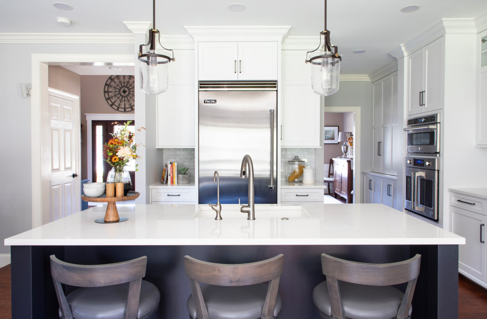 Example of a transitional medium tone wood floor kitchen design with an undermount sink, shaker cabinets, quartz countertops, gray backsplash, porcelain backsplash, stainless steel appliances, an island and white countertops