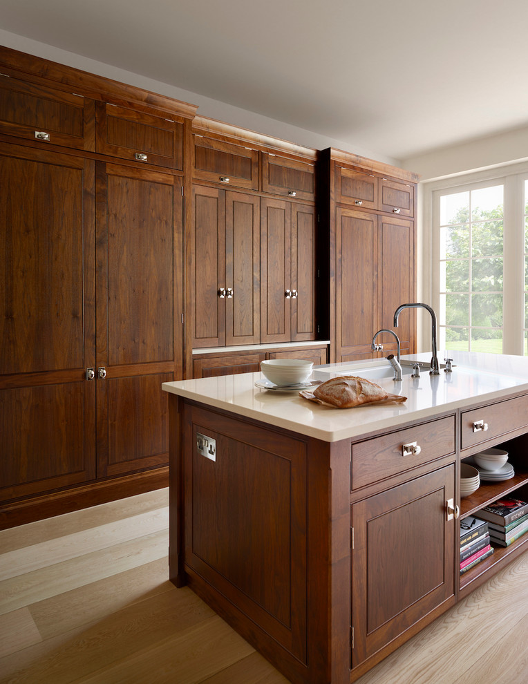 Photo of a large traditional galley eat-in kitchen in Essex with an undermount sink, raised-panel cabinets, dark wood cabinets, light hardwood floors, with island, solid surface benchtops and panelled appliances.