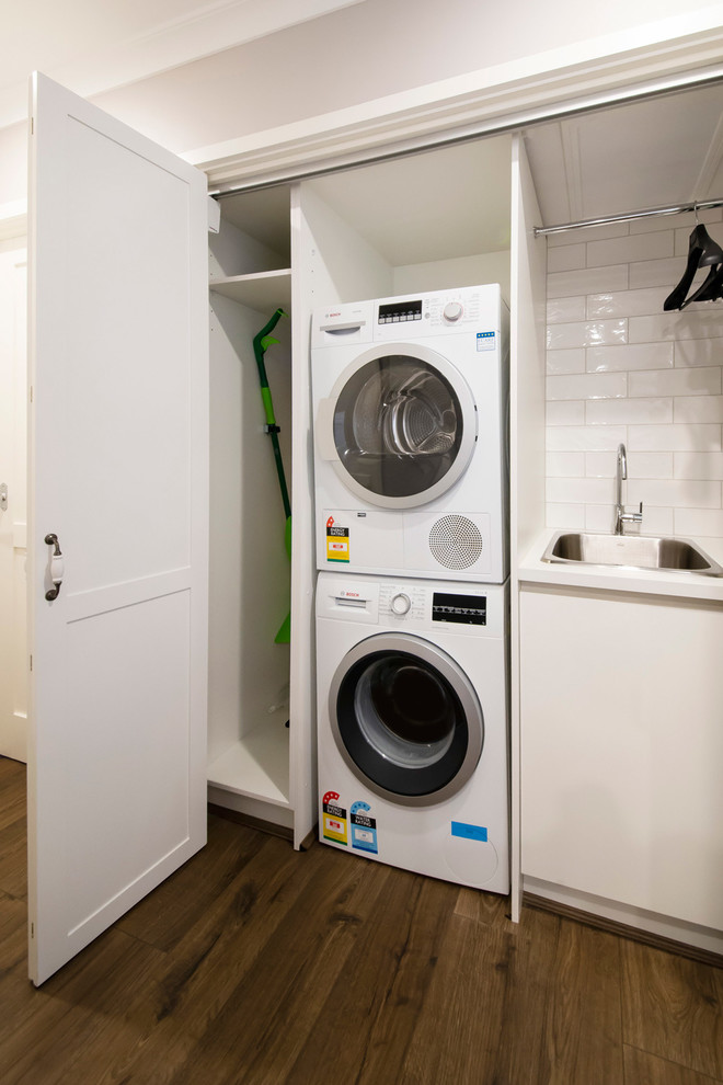 This is an example of a small traditional single-wall laundry cupboard in Melbourne with a drop-in sink, white cabinets, quartz benchtops, grey walls, laminate floors, a stacked washer and dryer, white benchtop and shaker cabinets.