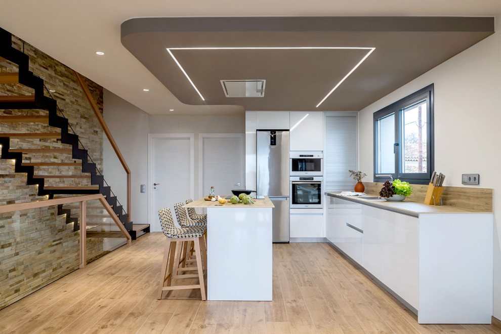 Modern inredning av ett beige beige kök med öppen planlösning, med släta luckor, vita skåp, träbänkskiva, rostfria vitvaror, ljust trägolv, en köksö och beiget golv