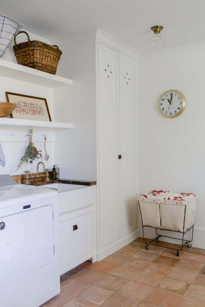 Inspiration for a large cottage single-wall terra-cotta tile and multicolored floor dedicated laundry room remodel in Dallas with a farmhouse sink, shaker cabinets, white cabinets, wood countertops, white walls, a side-by-side washer/dryer and brown countertops