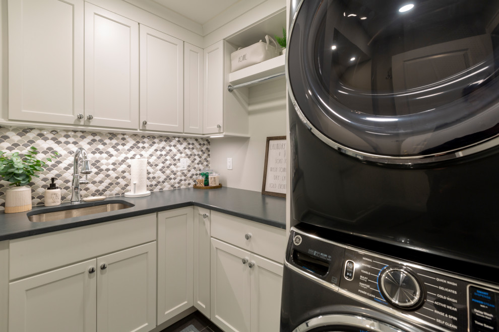 Photo of a mid-sized transitional l-shaped dedicated laundry room in New York with an undermount sink, shaker cabinets, white cabinets, quartz benchtops, multi-coloured splashback, mosaic tile splashback, beige walls, a stacked washer and dryer and black benchtop.