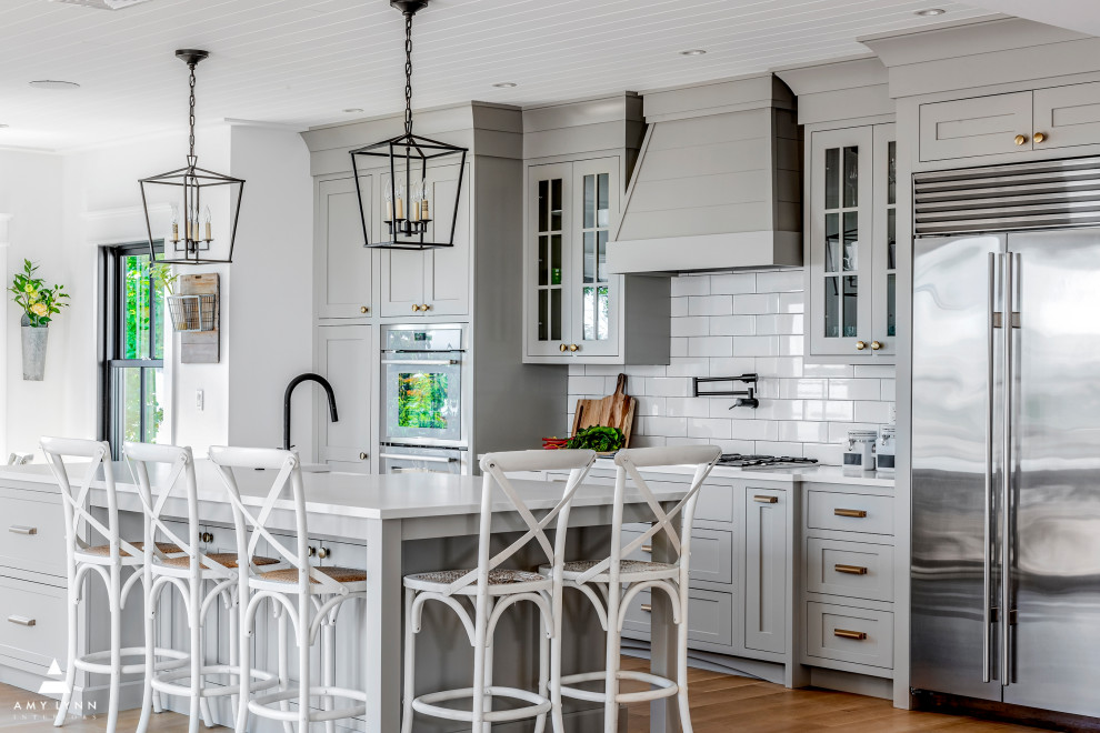 Photo of a large country u-shaped eat-in kitchen in Boston with a farmhouse sink, shaker cabinets, grey cabinets, quartz benchtops, white splashback, ceramic splashback, stainless steel appliances, light hardwood floors, with island, brown floor, white benchtop and timber.
