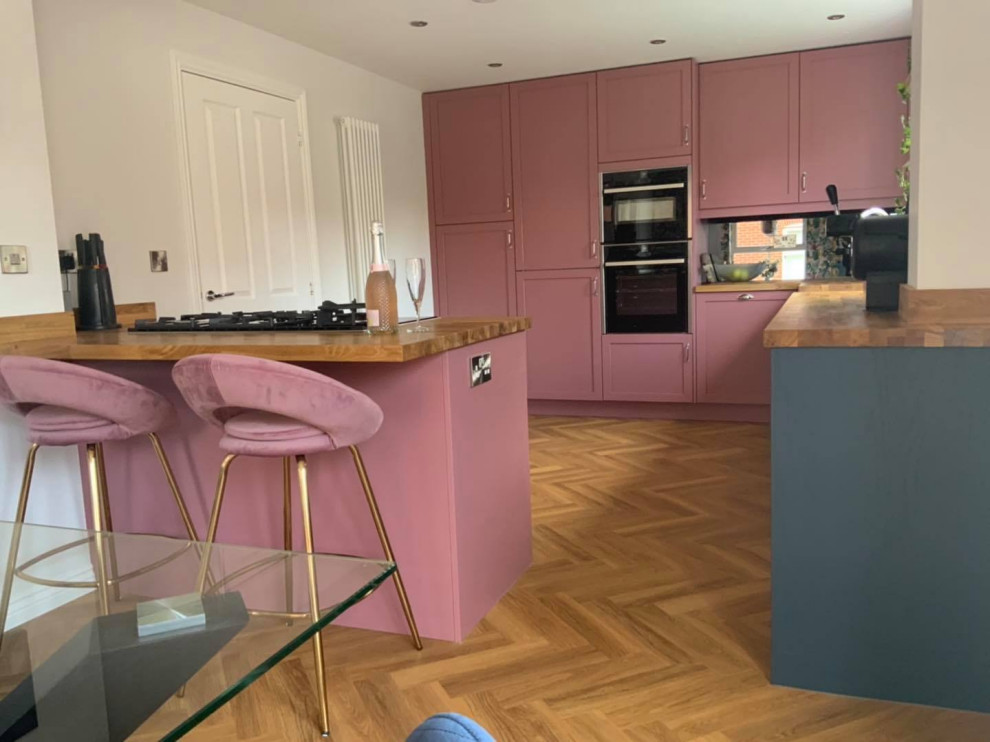 Inspiration for a mid-sized eclectic l-shaped eat-in kitchen in Buckinghamshire with a farmhouse sink, shaker cabinets, pink cabinets, wood benchtops, metallic splashback, mirror splashback, panelled appliances, vinyl floors, a peninsula, brown floor and brown benchtop.