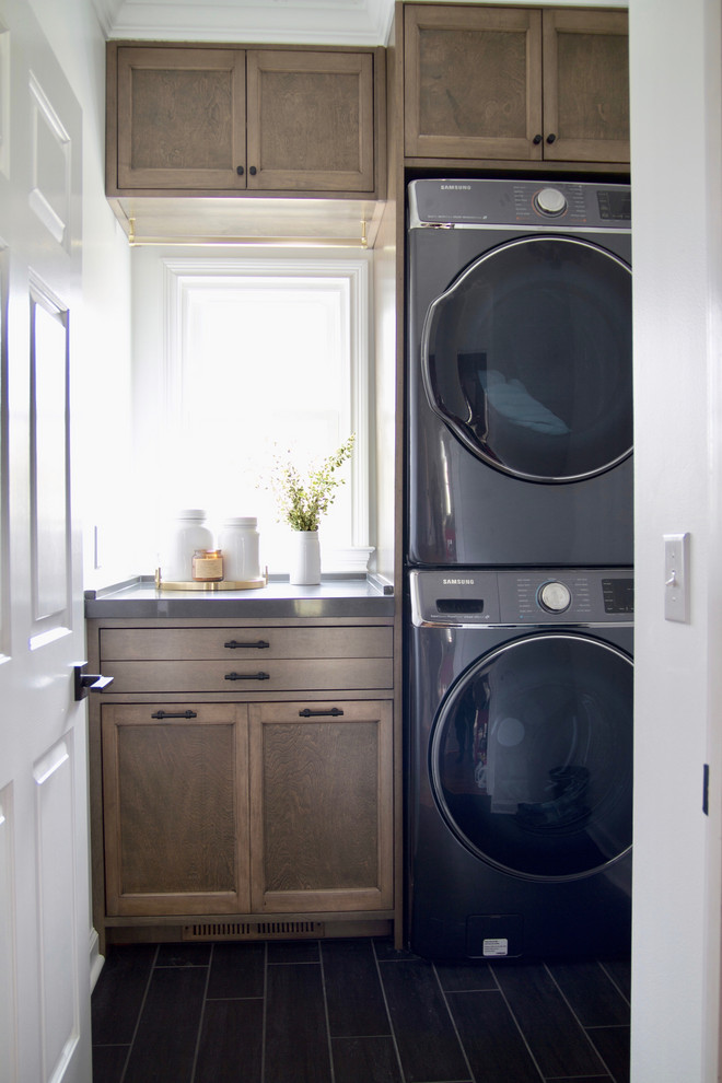 Small transitional single-wall dedicated laundry room with shaker cabinets, medium wood cabinets, quartz benchtops, white walls, porcelain floors, a stacked washer and dryer, black floor and grey benchtop.