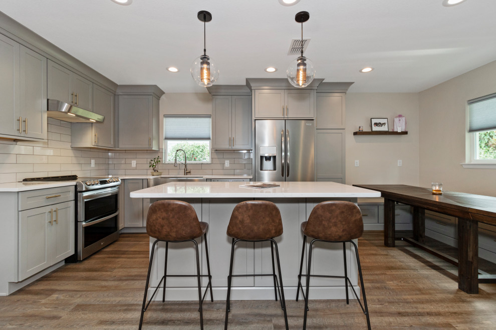 Photo of a large modern l-shaped open plan kitchen in Tampa with a double-bowl sink, shaker cabinets, grey cabinets, quartz benchtops, white splashback, subway tile splashback, stainless steel appliances, laminate floors, with island, brown floor and white benchtop.