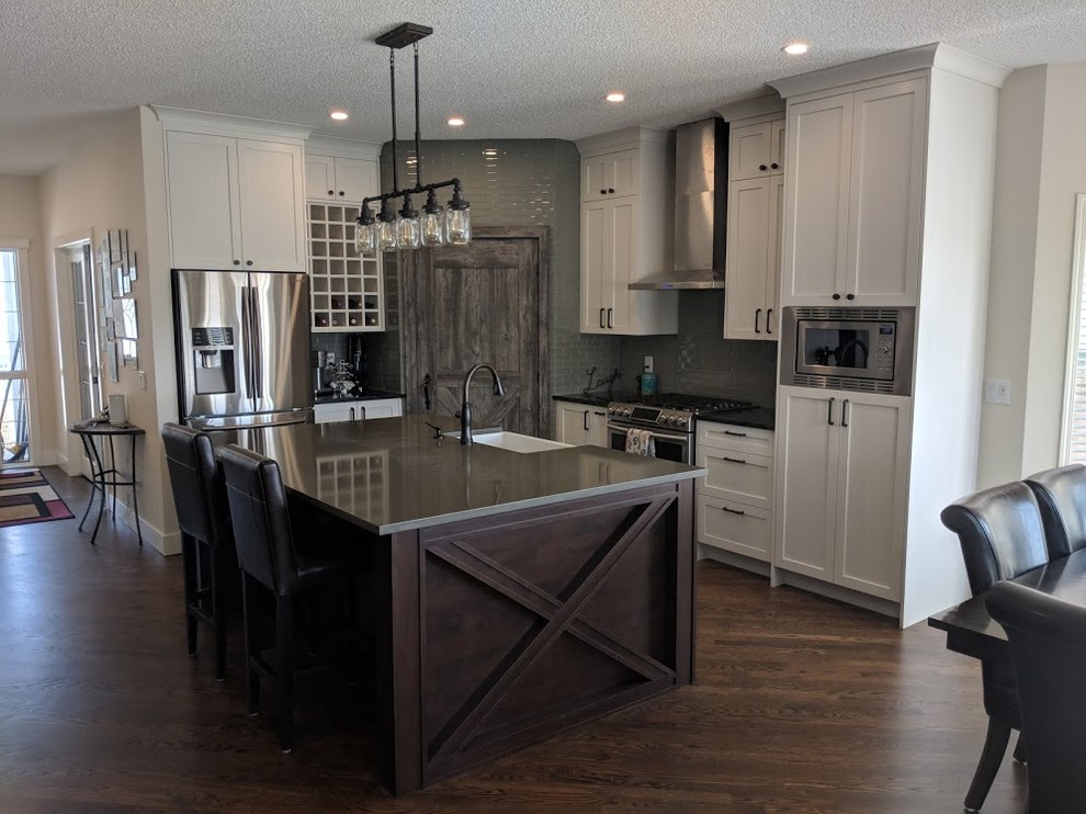 Inspiration for a mid-sized contemporary l-shaped eat-in kitchen in Calgary with a farmhouse sink, shaker cabinets, white cabinets, solid surface benchtops, grey splashback, subway tile splashback, stainless steel appliances, dark hardwood floors, with island, brown floor and grey benchtop.