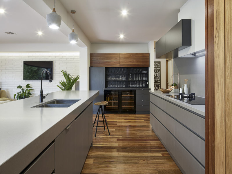 This is an example of a mid-sized modern galley open plan kitchen in Melbourne with medium hardwood floors, brown floor, a drop-in sink, flat-panel cabinets, grey cabinets, limestone benchtops, grey splashback, ceramic splashback, black appliances, with island and white benchtop.