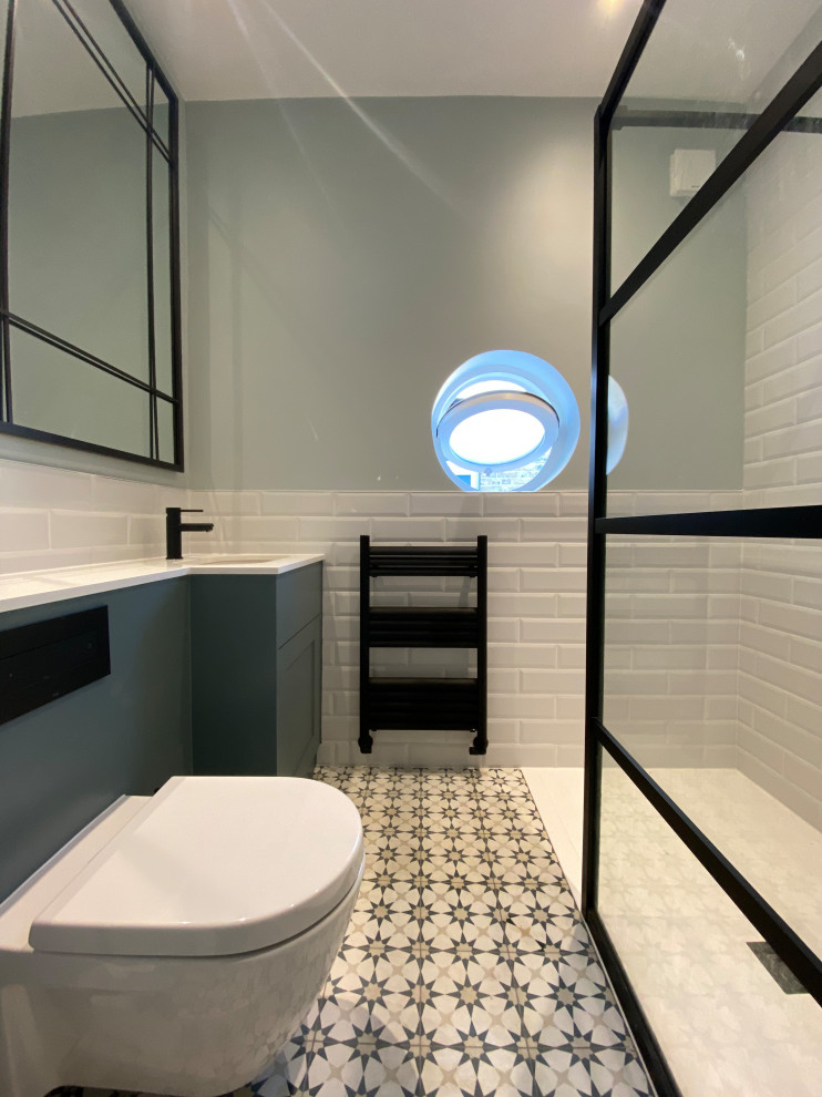 This is an example of a mid-sized contemporary kids bathroom in London with shaker cabinets, blue cabinets, a drop-in tub, an open shower, a wall-mount toilet, white tile, ceramic tile, white walls, ceramic floors, a drop-in sink, quartzite benchtops, multi-coloured floor, an open shower, white benchtops, a niche, a single vanity and a built-in vanity.
