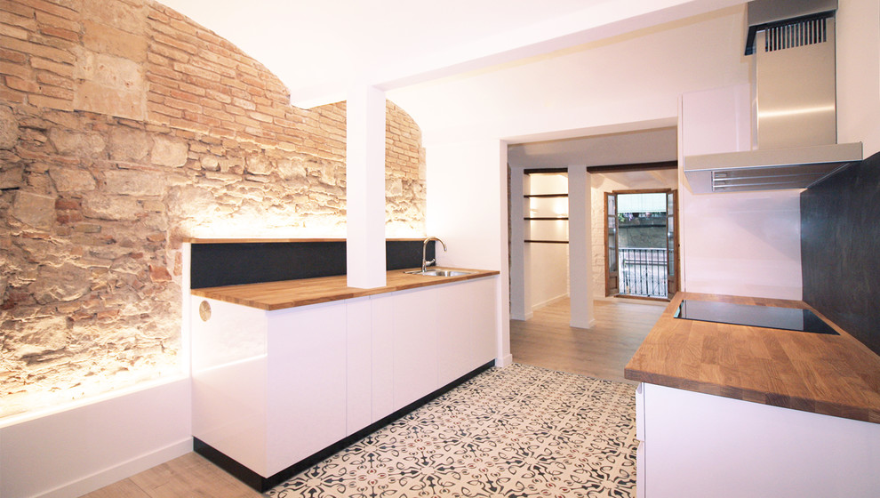 Photo of a mid-sized mediterranean single-wall open plan kitchen in Barcelona with an undermount sink, wood benchtops, black splashback, slate splashback, panelled appliances, cement tiles, multi-coloured floor, flat-panel cabinets, white cabinets, no island and brown benchtop.