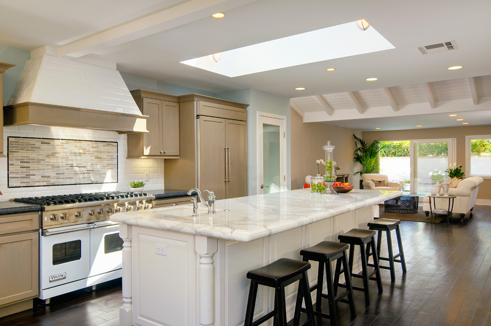 This is an example of a mid-sized traditional l-shaped open plan kitchen in Orange County with white appliances, marble benchtops, a drop-in sink, recessed-panel cabinets, white cabinets, white splashback, medium hardwood floors and with island.