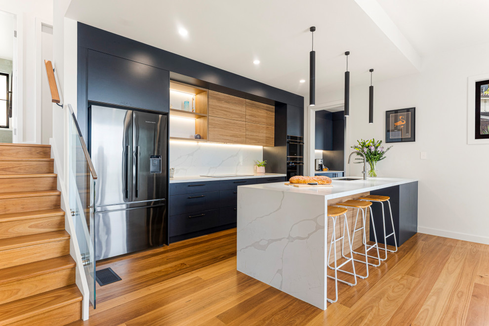 This is an example of a contemporary galley eat-in kitchen in Central Coast with an undermount sink, flat-panel cabinets, quartz benchtops, white splashback, engineered quartz splashback, black appliances, medium hardwood floors, with island, brown floor, white benchtop and black cabinets.