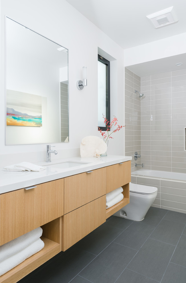 This is an example of a mid-sized beach style kids bathroom in Orange County with flat-panel cabinets, light wood cabinets, an alcove shower, gray tile, white walls, an undermount sink, engineered quartz benchtops, an alcove tub, a one-piece toilet, subway tile and ceramic floors.