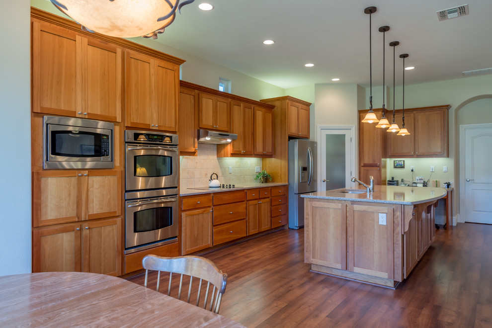 This is an example of a large traditional l-shaped eat-in kitchen in Sacramento with an undermount sink, shaker cabinets, medium wood cabinets, granite benchtops, beige splashback, terra-cotta splashback, stainless steel appliances, vinyl floors, with island and brown floor.
