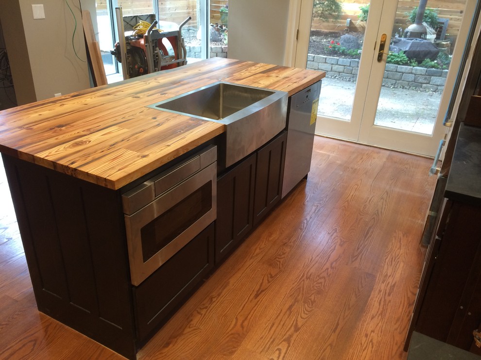 This is an example of a mid-sized arts and crafts l-shaped open plan kitchen in Philadelphia with shaker cabinets, black cabinets, wood benchtops, with island, a farmhouse sink, white splashback, porcelain splashback, stainless steel appliances, medium hardwood floors and brown floor.