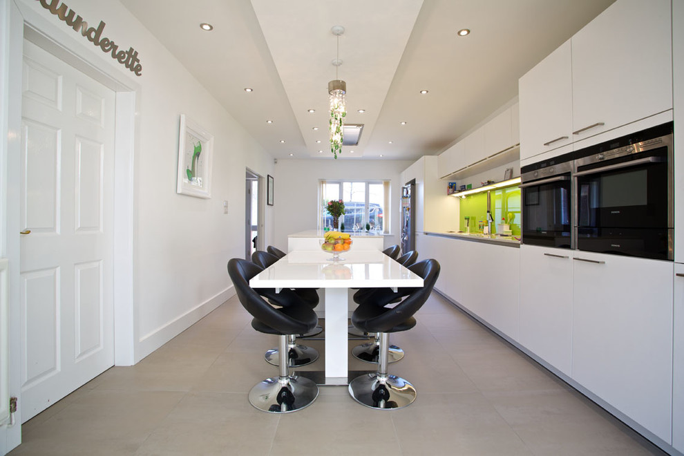Inspiration for a large contemporary galley open plan kitchen in Hampshire with flat-panel cabinets, white cabinets, green splashback, glass sheet splashback, stainless steel appliances, an integrated sink, solid surface benchtops, porcelain floors, with island and white benchtop.
