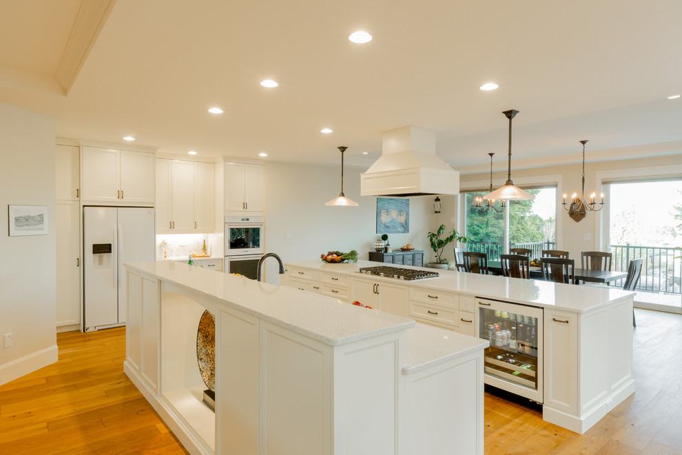 Photo of a large traditional open plan kitchen in Vancouver with a farmhouse sink, recessed-panel cabinets, white cabinets, quartzite benchtops, white splashback, white appliances, light hardwood floors and multiple islands.