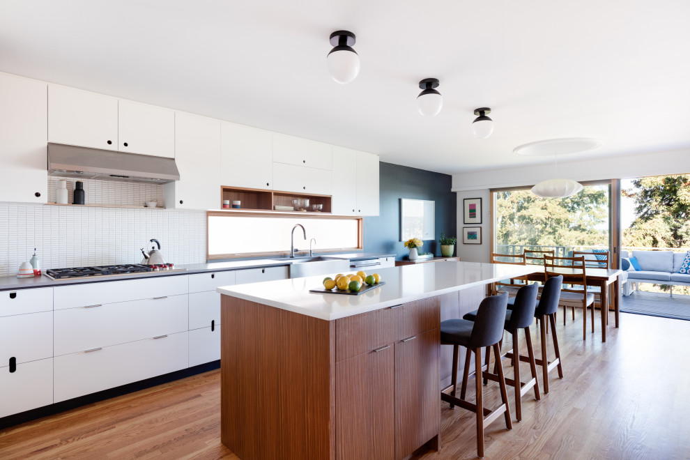 Design ideas for a mid-sized midcentury l-shaped eat-in kitchen in Seattle with flat-panel cabinets, white cabinets, solid surface benchtops, stainless steel appliances, medium hardwood floors, with island, white benchtop, a farmhouse sink, white splashback, mosaic tile splashback and brown floor.