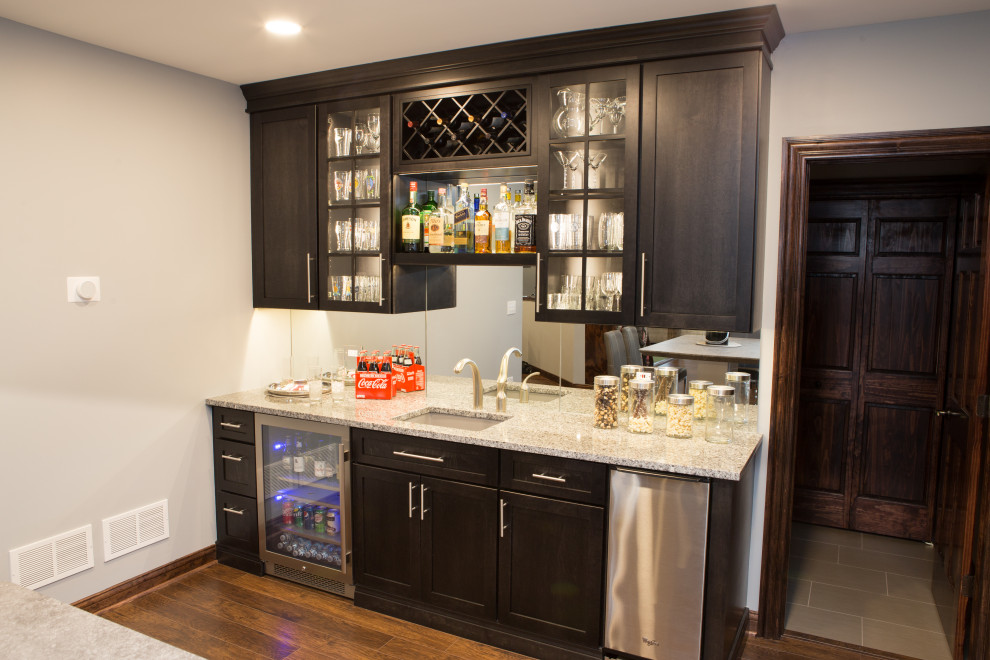 Design ideas for a large transitional single-wall wet bar in Chicago with an undermount sink, recessed-panel cabinets, granite benchtops, mirror splashback, laminate floors, brown floor, multi-coloured benchtop and grey cabinets.