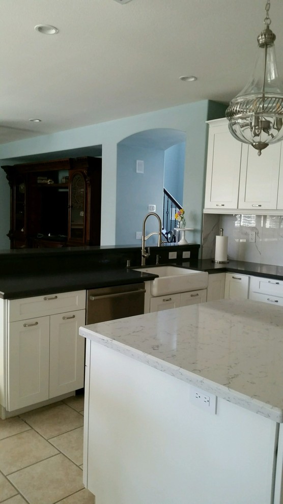 Photo of a mid-sized contemporary l-shaped eat-in kitchen in Los Angeles with a farmhouse sink, shaker cabinets, white cabinets, quartz benchtops, white splashback, subway tile splashback, stainless steel appliances, porcelain floors, with island, beige floor and black benchtop.