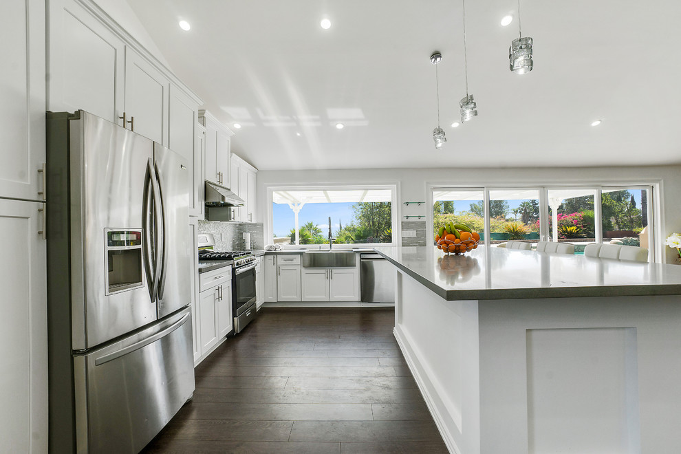 Photo of a large modern l-shaped eat-in kitchen in Seattle with a farmhouse sink, shaker cabinets, white cabinets, quartz benchtops, grey splashback, glass sheet splashback, stainless steel appliances, laminate floors, with island, brown floor and grey benchtop.