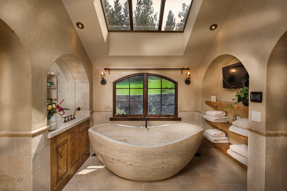 Photo of a large mediterranean master bathroom in Other with raised-panel cabinets, medium wood cabinets, beige tile, stone tile, a freestanding tub, beige walls, limestone floors and beige floor.