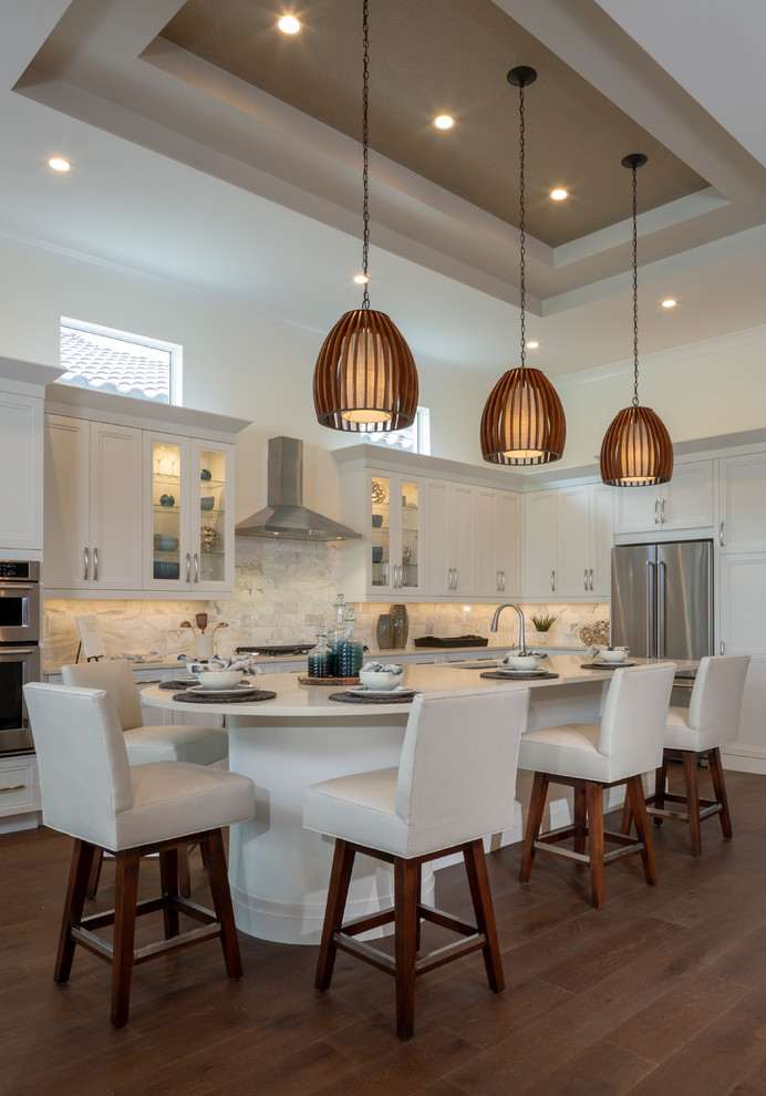 Transitional l-shaped kitchen in Miami with an undermount sink, recessed-panel cabinets, white cabinets, white splashback, stainless steel appliances, medium hardwood floors, with island, brown floor and white benchtop.