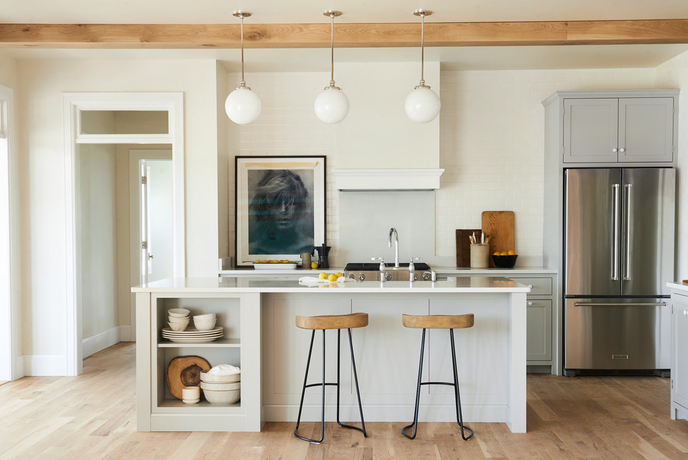 Example of a mid-sized transitional l-shaped light wood floor and brown floor open concept kitchen design with shaker cabinets, gray cabinets, white backsplash, subway tile backsplash, stainless steel appliances and an island
