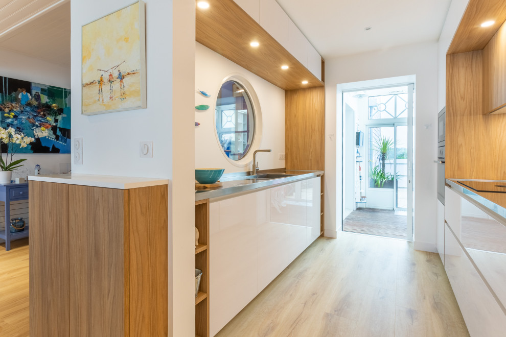 Photo of a small beach style galley open plan kitchen in Rennes with an integrated sink, beaded inset cabinets, white cabinets, stainless steel benchtops, mirror splashback, stainless steel appliances, light hardwood floors and timber.