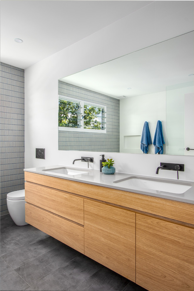 This is an example of a contemporary kids bathroom in Sydney with furniture-like cabinets, light wood cabinets, a freestanding tub, an open shower, a wall-mount toilet, blue tile, matchstick tile, white walls, cement tiles, an undermount sink, engineered quartz benchtops, grey floor, an open shower and grey benchtops.