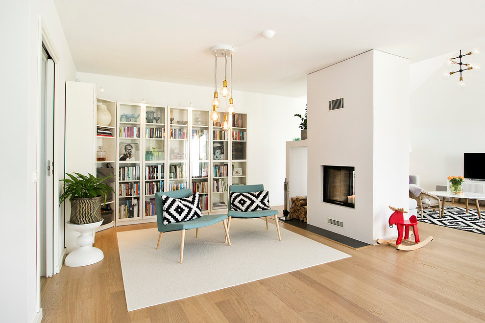 This is an example of a mid-sized scandinavian open concept living room in Malmo with a library, white walls, light hardwood floors, a two-sided fireplace and a freestanding tv.