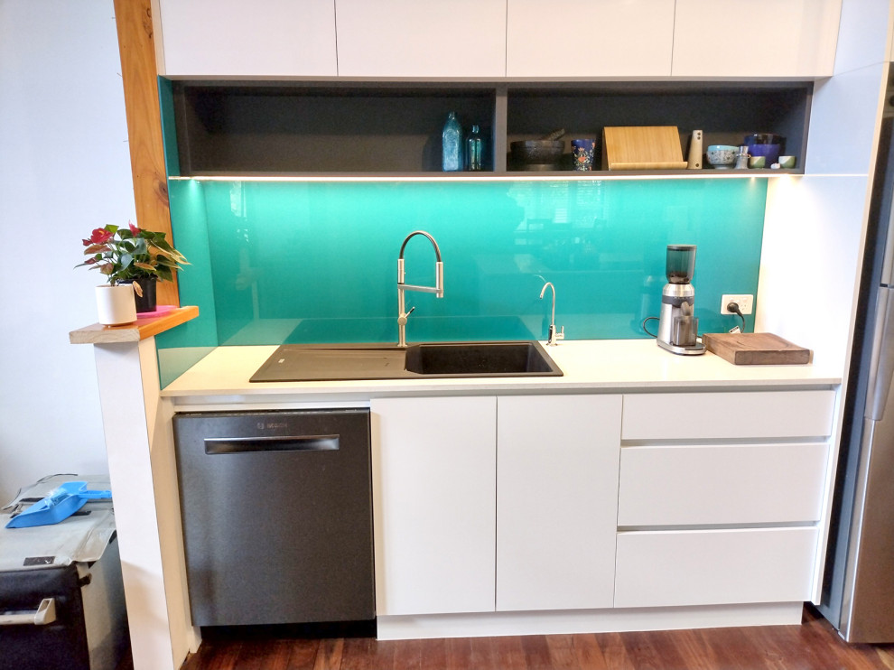 Photo of a mid-sized contemporary galley open plan kitchen in Perth with flat-panel cabinets, white cabinets, quartz benchtops, a peninsula, a drop-in sink, blue splashback, glass sheet splashback, black appliances, dark hardwood floors and white benchtop.