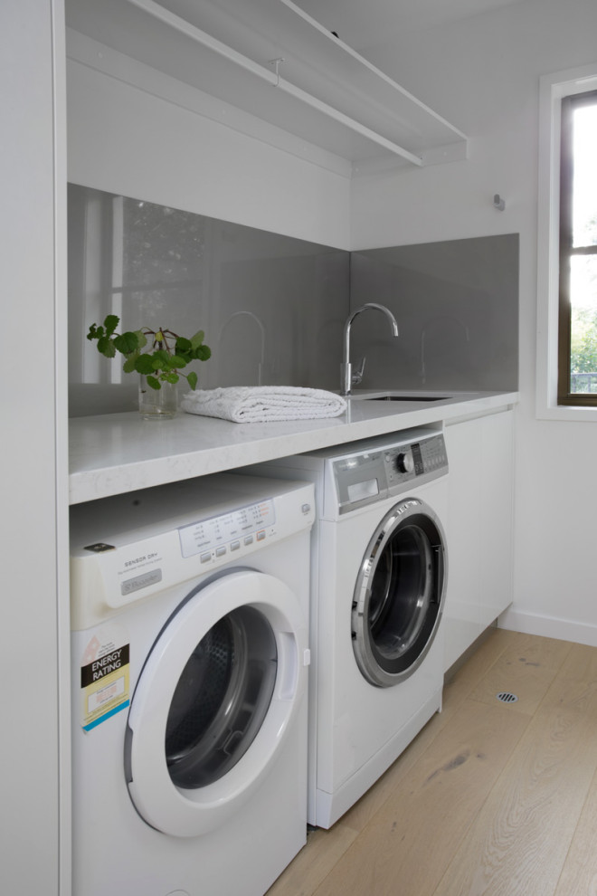 Photo of a small contemporary utility room in Brisbane with an undermount sink, white cabinets, quartz benchtops, metallic splashback, white walls, light hardwood floors, a side-by-side washer and dryer and white benchtop.