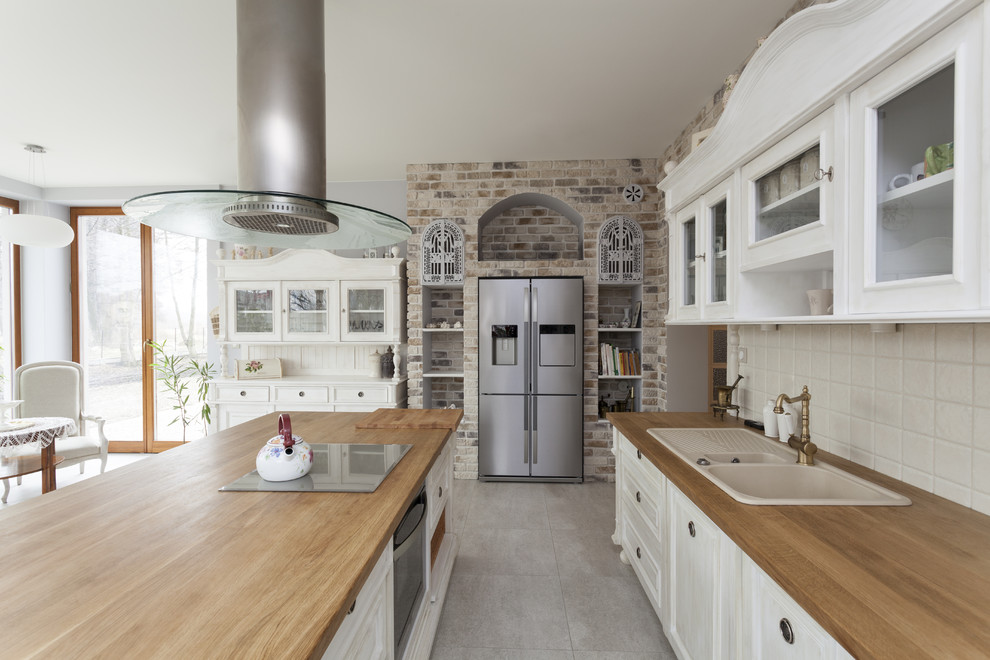 Large traditional l-shaped eat-in kitchen in Sydney with stainless steel appliances, with island, a drop-in sink, white cabinets, wood benchtops, white splashback, ceramic splashback, ceramic floors, raised-panel cabinets, brown floor and brown benchtop.