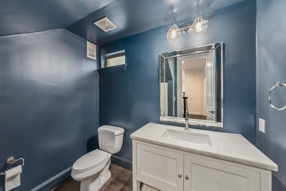 This is an example of a small transitional powder room in Houston with recessed-panel cabinets, white cabinets, quartzite benchtops, white benchtops, a one-piece toilet, blue walls, laminate floors, an undermount sink and brown floor.