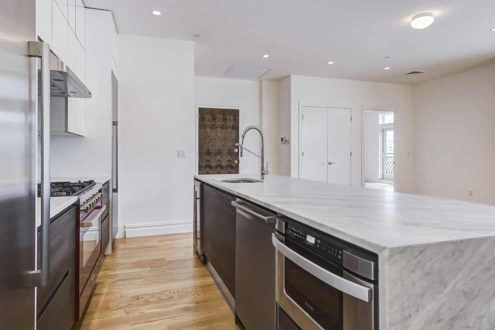 This is an example of a mid-sized modern single-wall open plan kitchen in New York with a drop-in sink, flat-panel cabinets, white cabinets, quartz benchtops, stone slab splashback, stainless steel appliances, light hardwood floors, brown floor, multi-coloured splashback and with island.