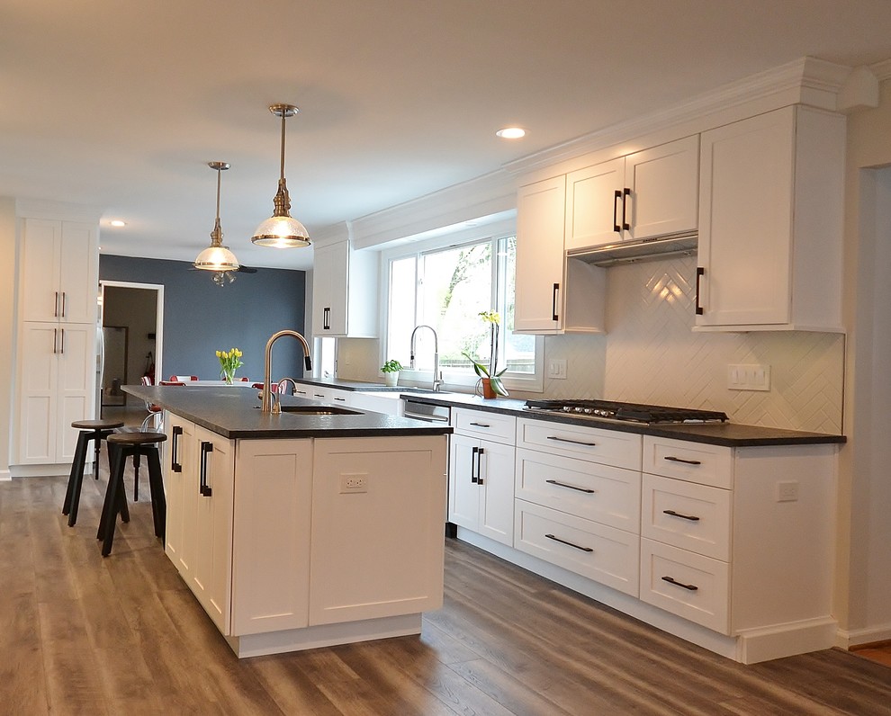 Photo of a large transitional l-shaped eat-in kitchen in Philadelphia with a farmhouse sink, recessed-panel cabinets, white cabinets, granite benchtops, white splashback, subway tile splashback, stainless steel appliances, vinyl floors, with island and brown floor.