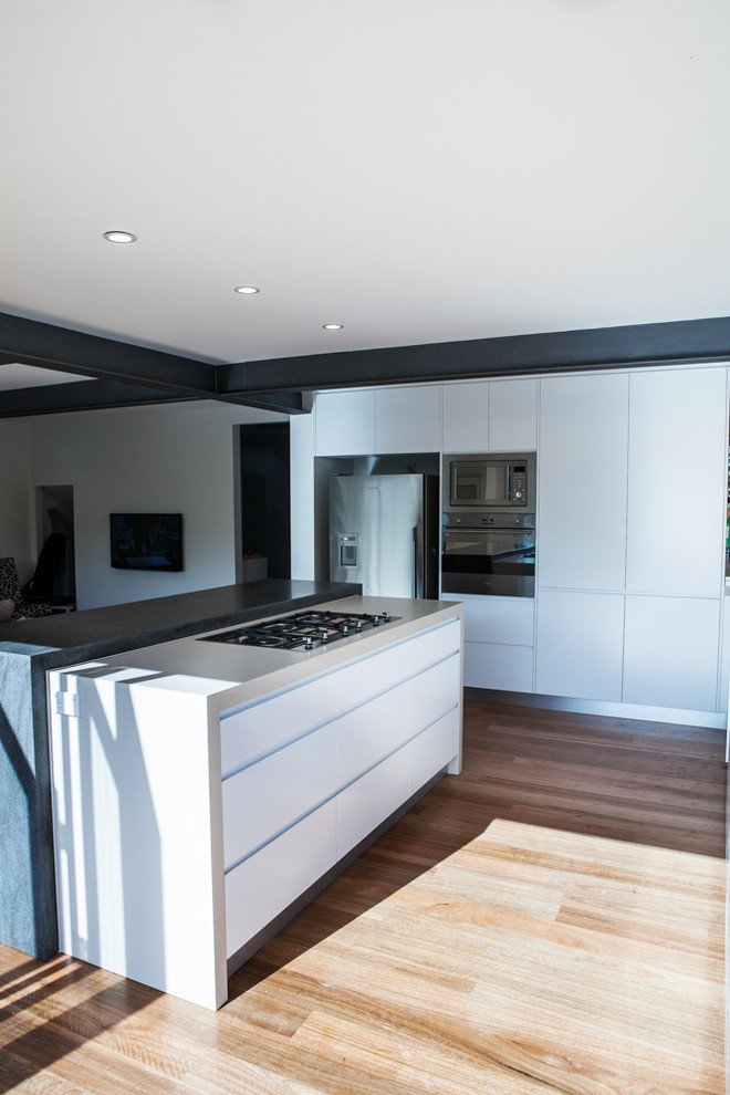 Large contemporary l-shaped open plan kitchen in Sydney with an undermount sink, flat-panel cabinets, white cabinets, solid surface benchtops, stainless steel appliances, medium hardwood floors and with island.