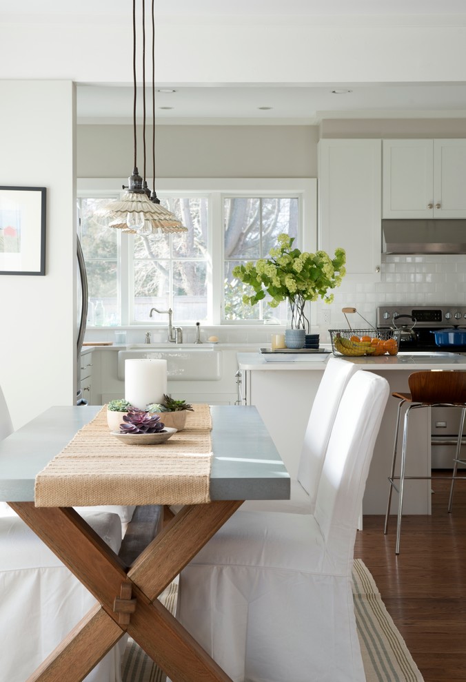This is an example of a mid-sized beach style l-shaped eat-in kitchen in New York with medium hardwood floors, a farmhouse sink, shaker cabinets, white cabinets, solid surface benchtops, white splashback, ceramic splashback, stainless steel appliances, with island and brown floor.