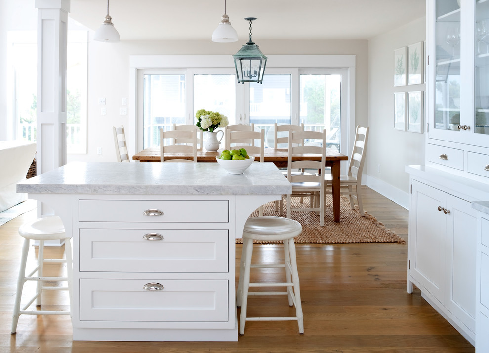 This is an example of a beach style open plan kitchen in Other with a farmhouse sink, shaker cabinets, white cabinets, marble benchtops, white splashback, subway tile splashback, white appliances, light hardwood floors and with island.
