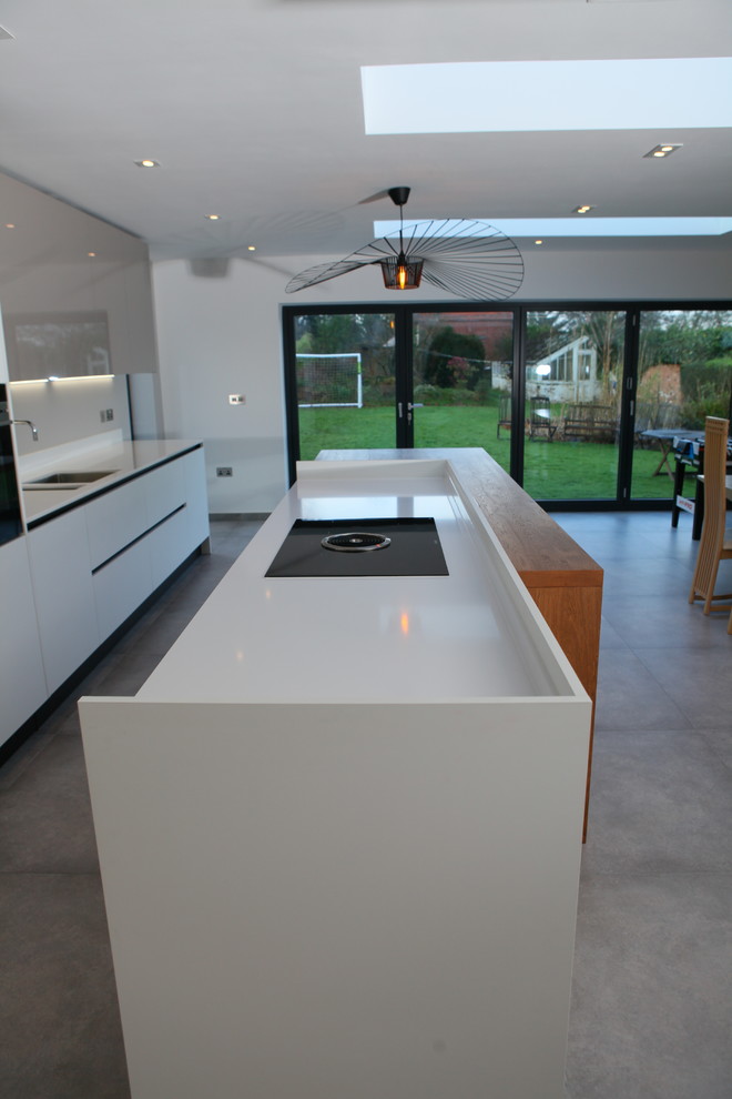 Large contemporary single-wall open plan kitchen in West Midlands with a drop-in sink, flat-panel cabinets, white cabinets, solid surface benchtops, black appliances, cement tiles, with island and grey floor.