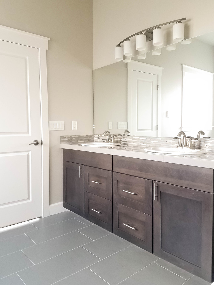 Photo of a mid-sized transitional master bathroom in Seattle with shaker cabinets, brown cabinets, a drop-in tub, a one-piece toilet, gray tile, mosaic tile, grey walls, porcelain floors, a drop-in sink, tile benchtops, grey floor and a hinged shower door.