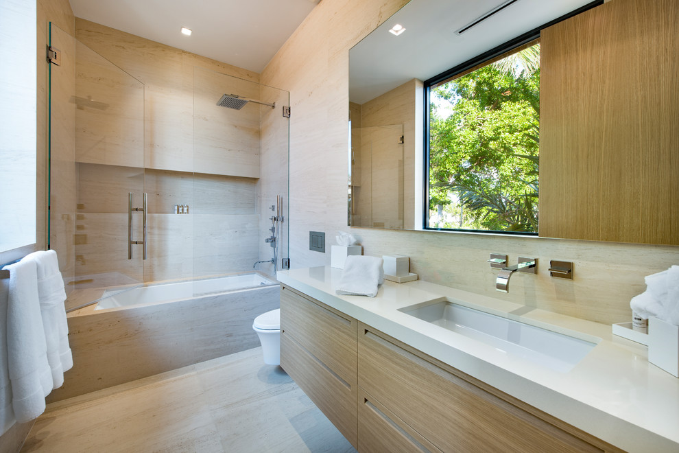 Design ideas for a mid-sized modern bathroom in Miami with flat-panel cabinets, beige cabinets, a freestanding tub, a shower/bathtub combo, a wall-mount toilet, beige tile, stone tile, beige walls, light hardwood floors, an undermount sink, marble benchtops, a hinged shower door and white benchtops.