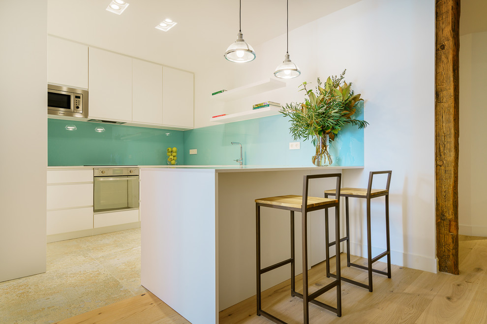 This is an example of a contemporary u-shaped kitchen in Madrid with flat-panel cabinets, white cabinets, blue splashback, glass sheet splashback, stainless steel appliances, light hardwood floors, a peninsula and grey benchtop.