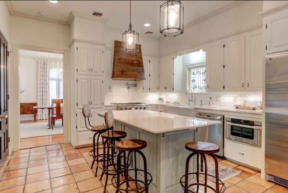 This is an example of a large country kitchen in Dallas with a farmhouse sink, raised-panel cabinets, white cabinets, quartzite benchtops, white splashback, subway tile splashback, stainless steel appliances, terra-cotta floors, with island and white benchtop.