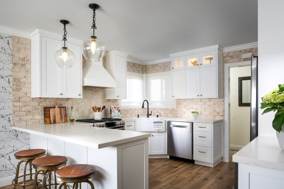 This is an example of a mid-sized transitional u-shaped eat-in kitchen in Manchester with a farmhouse sink, shaker cabinets, white cabinets, solid surface benchtops, beige splashback, brick splashback, stainless steel appliances, medium hardwood floors, a peninsula, brown floor and beige benchtop.